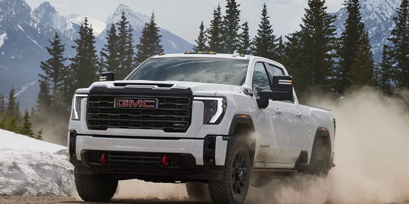 A white 2026 GMC Sierra 2500 heavy-duty truck showcasing rugged capability is driving rapidly up a snow-covered mountain road.