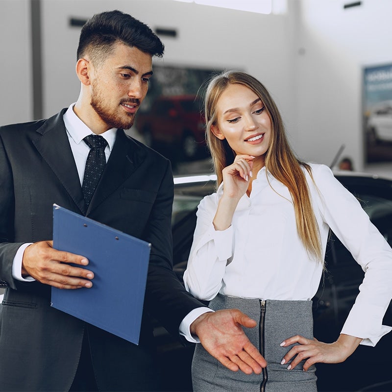 A GMC sales person in a dark suit & tie and a woman with long blonde hair are smiling as he explains the terms of the sale of a vehicle to her.